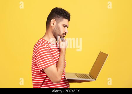 Profile portrait thoughtful pensive man with beard in striped t-shirt holding laptop and rubbing chin looking at display, thinking over new business p Stock Photo