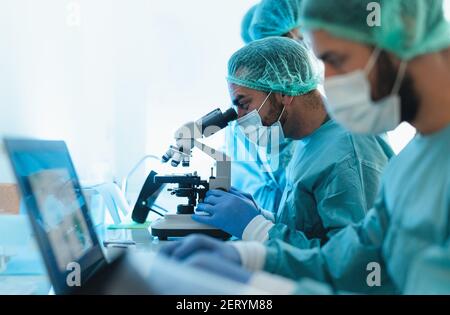 Doctors wearing personal protective equipment working in laboratory using computer and examining coronavirus through microscope Stock Photo