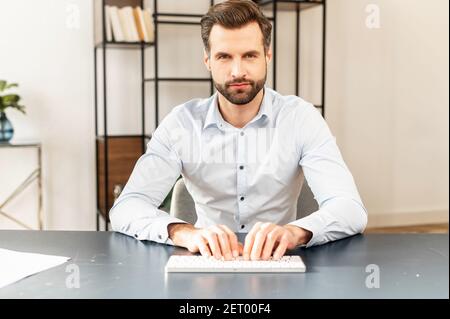Serious concentrated young bearded entrepreneur wearing smart casual clothes, sitting at the desk, typing on keyboard, businessman professional is fee Stock Photo