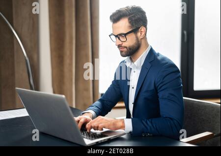 Serious concentrated determined young bearded office worker in business casual clothes, sitting at the desk, typing on laptop, businessman professiona Stock Photo