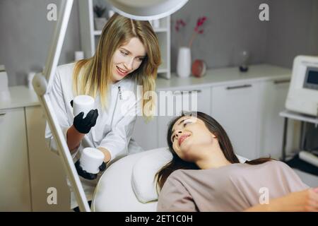 Consultation in cosmetology clinic. Female beauty doctor talking with patient and showing her what she's going to use, cream or mask. Stock Photo