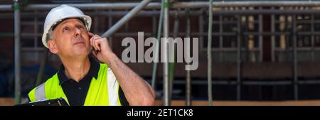 Panorama thoughtful male builder foreman, construction worker, contractor or architect on building site holding black clipboard and talking on his cel Stock Photo