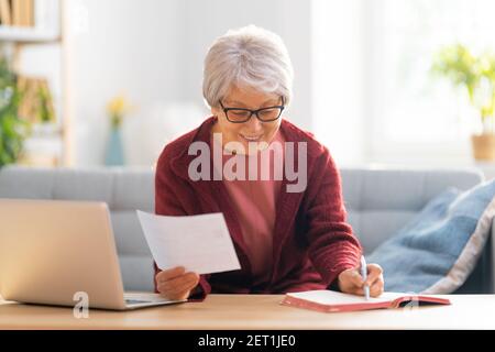 Elderly woman, sitting on the sofa with a paper receipt in her hands, are calculating expenses, managing the family budget. Stock Photo