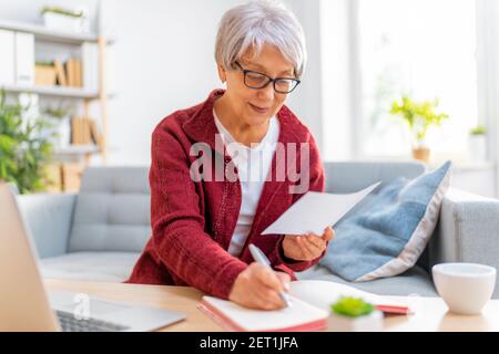 Elderly woman, sitting on the sofa with a paper receipt in her hands, are calculating expenses, managing the family budget. Stock Photo