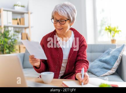 Elderly woman, sitting on the sofa with a paper receipt in her hands, are calculating expenses, managing the family budget. Stock Photo