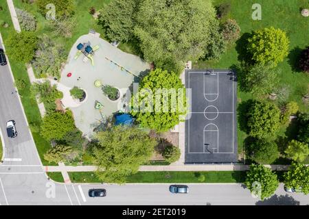 An aerial drone view of a local park with playground equipment, a lot of grass and trees, and a full basketball court. Stock Photo