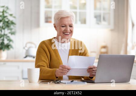 Positive grandmother sitting in kitchen, reading letter Stock Photo