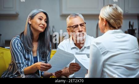 elderly interracial couple holding documents near insurance agent on blurred foreground Stock Photo