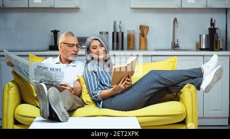 smiling asian woman reading book near senior husband with newspaper Stock Photo