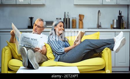 elderly interracial couple reading book and newspaper on sofa at home Stock Photo