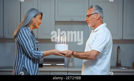 side view of smiling senior man presenting birthday cake to joyful asian wife Stock Photo