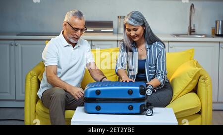 senior interracial couple sitting on couch at home near suitcase Stock Photo