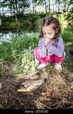Girl crouching by a riverbank looking at a freshly caught fish, Poland Stock Photo