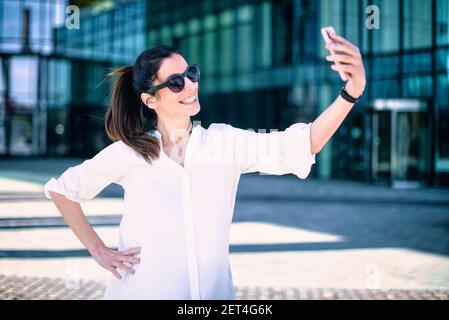 Outdoor shot of happy woman taking self portrait with her mobile phone while standing outdoor. Stock Photo