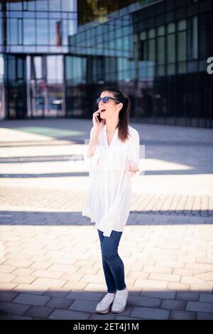 Full length shot of attractive businesswoman wearing white shirt and sunglasses while talking with somebody on her mobile phone outdoor. Stock Photo