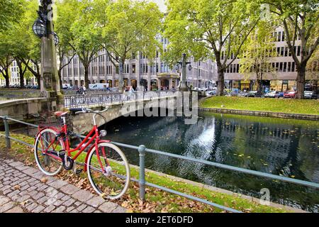 Königsallee is not only a famous shopping boulevard, but also has wonderful green areas. Romantic scenery with red bike at the city canal 'Kö-Graben'. Stock Photo