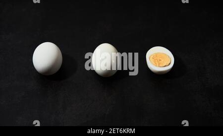 Three chicken eggs lie on black kitchen table side view from below. Whole egg in white shell, peeled and cut in half so that you can see boiled yolk. Stock Photo