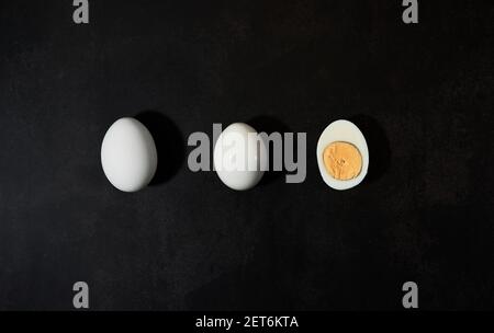Three chicken eggs lie on black kitchen table top view. Whole egg in white shell, peeled and cut in half so that you can see boiled yolk. Minimalistic Stock Photo