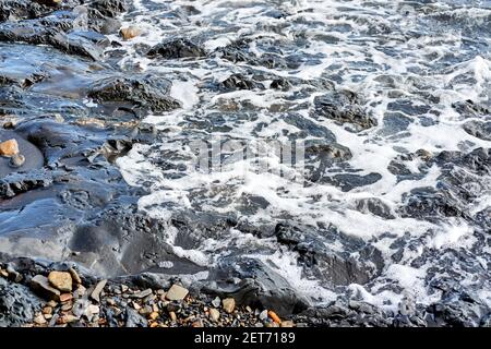 Small wave rolls onto shore and splash of foam flies in different directions. Beautiful marine minimalist screensaver. Sea waves break on shingle beac Stock Photo