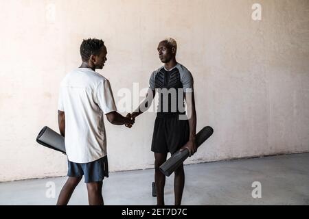 Two young african american men with exercise mats shaking hands while standing over wall indoors Stock Photo