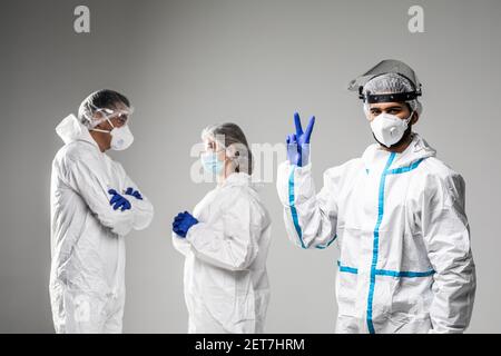 Male doctor with v-sign gesture standing in front of collegues that have informal meeting wearing in hazard on white background Stock Photo