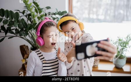 Two small girls sisters with headphones indoors at home, grimacing when taking selfie. Stock Photo