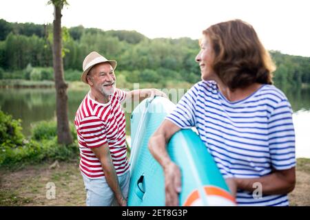 Senior man carrying paddleboard by lake in summer, talking. Stock Photo