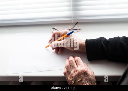 Hands of old woman with pen writing on piece of paper,  next to the window. Stock Photo
