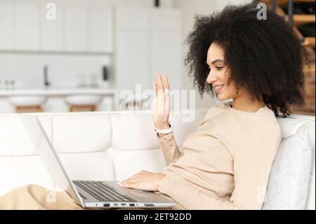 Video call concept. Cheerful African American female blogger with voluminous dark hair, waving hand while having video call on laptop from home, laying on the sofa, freelance work anytime, anywhere Stock Photo