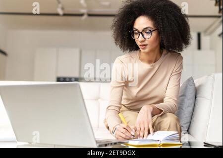 Elegant young adult female psychologist, working at home, writing down in her diary, sitting at the laptop, consulting the client online, talking, feeling confidently, love her work, support others Stock Photo