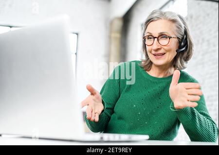 Senior gray-haired female employee in green jumper and glasses is sitting at the desk, wearing wireless headset, looking at the laptop screen, gesturing and talking online, video-calling with client Stock Photo