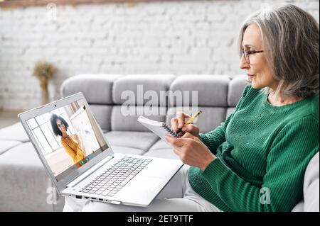 Attentive mature gray-haired businesswoman in glasses sitting on the couch at home, studying, taking virtual class, having online meeting on a laptop, taking notes and listening to female teacher Stock Photo