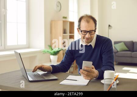 Mature man using smart app on mobile phone while sitting at office desk with laptop Stock Photo