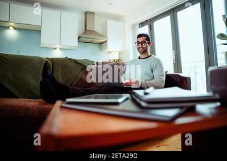 Mixed race business man working from home typing on laptop sitting on comfy couch Stock Photo