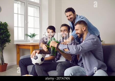 Smiling excited men friends clinking bottles with beer during watching television football match Stock Photo