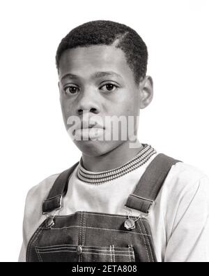 1970s PORTRAIT OF PRE-TEEN AFRICAN-AMERICAN BOY WEARING DENIM BIB OVERALLS SERIOUS FACIAL EXPRESSION LOOKING AT CAMERA  - b25648 HAR001 HARS SADNESS EYE CONTACT HEAD AND SHOULDERS AFRICAN-AMERICANS AFRICAN-AMERICAN BLACK ETHNICITY TEENAGED SINCERE SOLEMN BIB FOCUSED INTENSE JUVENILES PRE-TEEN PRE-TEEN BOY BLACK AND WHITE CAREFUL EARNEST HAR001 INTENT OLD FASHIONED AFRICAN AMERICANS Stock Photo