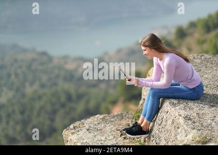 Full bofy side view portrait of a happy teen checking smart phone in a cliff Stock Photo
