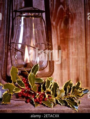 1970s ANTIQUE LANTERN WITH FLAME LIT ON WOOD BACKGROUND WITH SPRIG OF CHRISTMAS GREEN HOLLY WITH RED BERRIES - kx8762 HAR001 HARS LANTERNS GREENS HAR001 OLD FASHIONED RUSTY Stock Photo