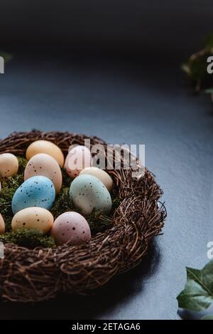 A wicker nest with moss and colored Easter eggs and green leaves on dark background. Dark mood style. Happy Easter holiday. Vertical card. Selective Stock Photo