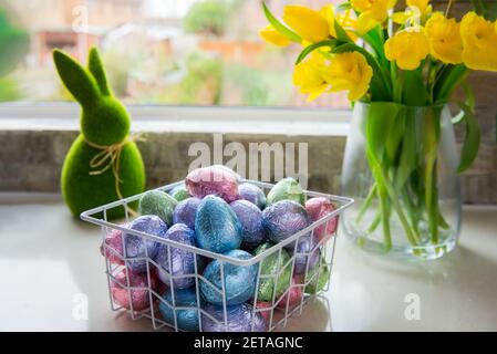Metal basket with chocolate easter eggs in color foil, bouquet of yellow tulips and daffodils flower and Easter green bunny on white kitchen table Stock Photo