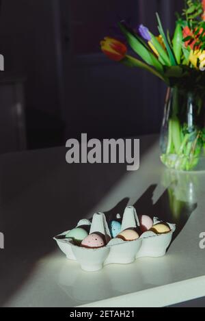 Easter celebration minimalism concept. Pastel colors painted eggs in a cardboard box and fresh bouquet of spring flowers on white kitchen table Stock Photo