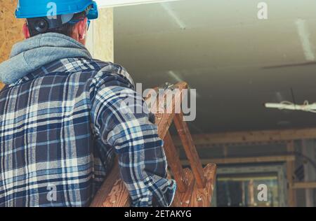 Construction Worker with Wooden Ladder in His Hands Moving Equipment to Another Room Inside Newly Constructed House. Stock Photo