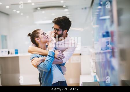 Young handsome joyful man is holding his girlfriend hugged from back as she is explaining him something.