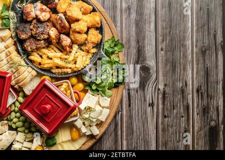 Top view on the wooden board plate on the table with various food appetizer catering french fries potato chips barbecue cheese bread and greens with c Stock Photo