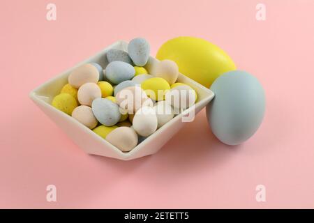 Pastel colored candy Easter eggs in white dessert bowl with two painted Easter eggs on pink background Stock Photo