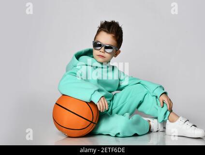 Cool kid boy in modern green, mint color sportswear hoodie, pants and white sneakers sits with basketball looking back