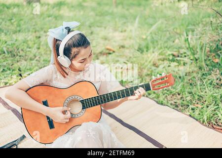 Girl wearing headphone to listen music and playing guitar in the forest. Music hobby and picnic concept. Stock Photo