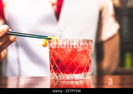 Close up shot of bartender hands preparing negroni cocktail with grapefruit. He is putting some essence from grapefruit skin into the cocktail glass on counter Stock Photo
