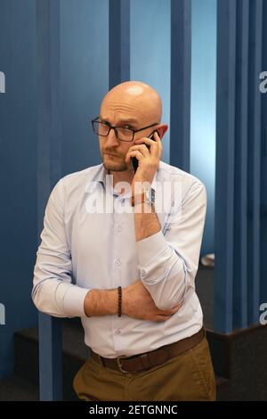 Adult bald man with glasses looks at camera, calls on mobile phone. Serious businessman solves problems Stock Photo
