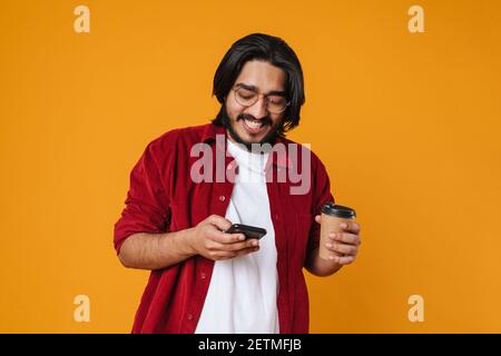 Happy handsome young man using his cellphone and holding cup of coffee isolated on yellow background Stock Photo